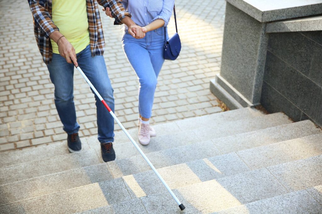 Uomo maturo con una disabilità visiva sale le scale accompagnato da una donna. Grazie alla riforma della disabilità del 2024 si va sempre più incontro all'integrazione totale delle persone con disabilità