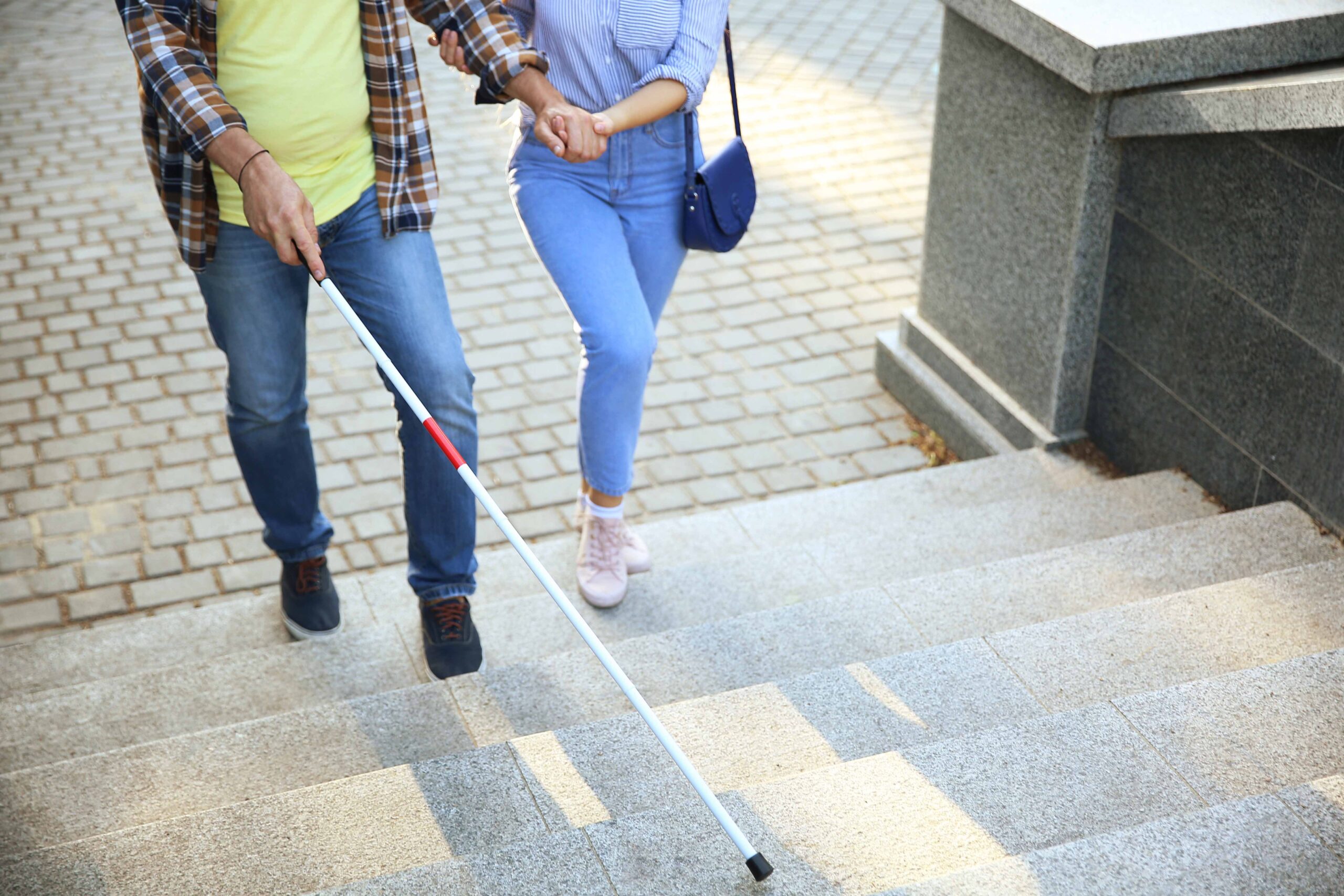 Uomo maturo con una disabilità visiva sale le scale accompagnato da una donna. Grazie alla riforma della disabilità del 2024 si va sempre più incontro all'integrazione totale delle persone con disabilità